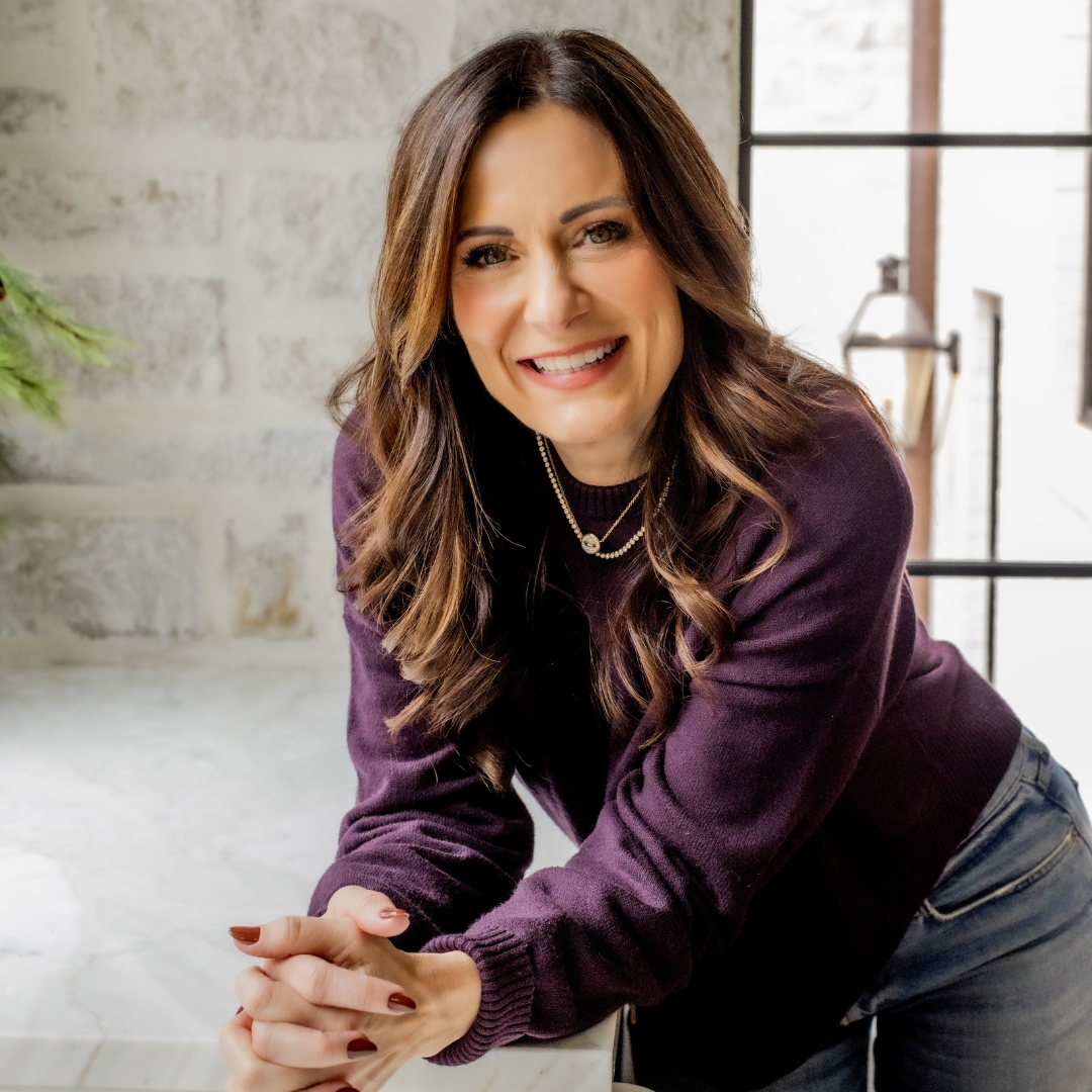Lysa TerKeurst wearing a purple blouse and leaning over a kitchen counter